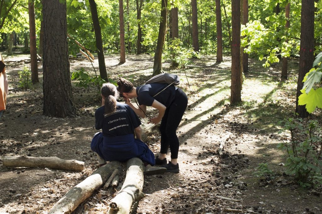 Das Foto ist in einem Wald aufgenommen worden. Zwischen dünnen Baumstämmen sitzt ein Mann mit Haarzopf auf einem liegenden Baumstamm. Er ist mit dem Rücken zum Betrachter des Bilder gedreht. Nebem ihm steht eine junge Frau mit Rucksack. Sie hat sich zu ihm hinuntergebeugt, ihm etwas zu zeigen. Sie schaut durch eine Kamera in die Richtung, in die auch der Mann blickt. Wir sehen beide Peronsen im Halbprofil.