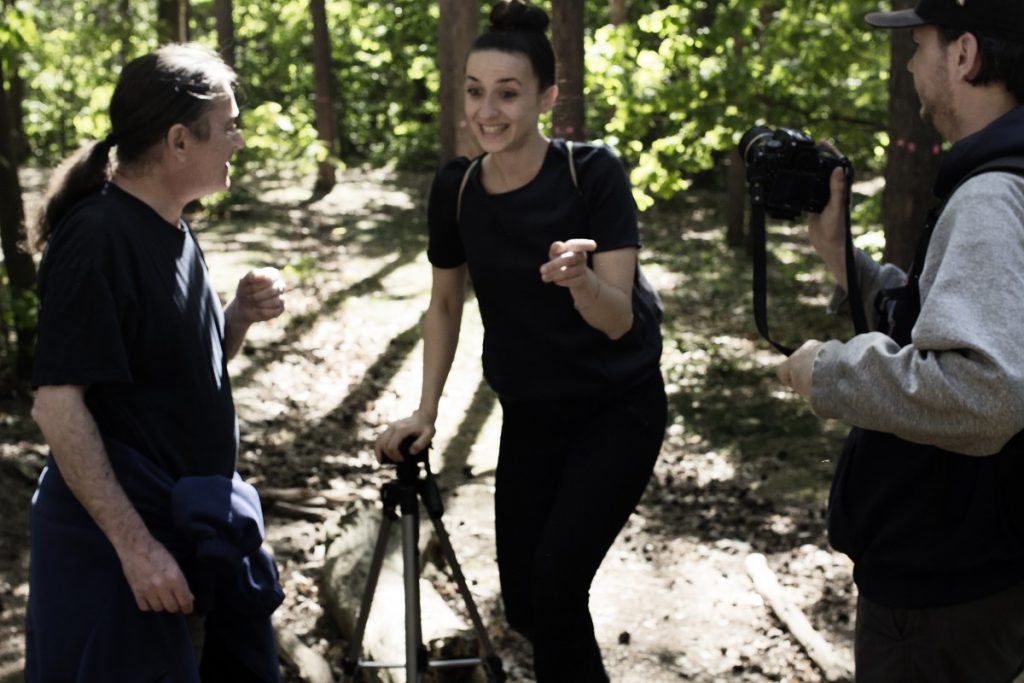 Auf dem Foto sind drei Personen zu sehen. In der Mitte steht eine junge Frau, sie blickt nach links unten und zeigt mit dem Zeigefinger in Richtung eines jungen Mannes, der rechts am Rand zu sehen ist. Er hält eine Kamera auf die junge Frau gerichtet. Es scheint, als würde er gleich den Auslöser drücken. Am linken Bildrand steht ein Mann mit Pferdeschwanz. Seiner Handhaltung und seinem Gesicht nach zu urteilen, spricht er gerade mit der jungen Frau oder hört ihr aufmerksam zu. Die junge Frau stützt sich mit der rechten Hand auf ein Stativ. Im Hintergrund sind Bäume zu sehen und Laub, welches hellgrün vom Sonnenlicht durchschienen wird.
