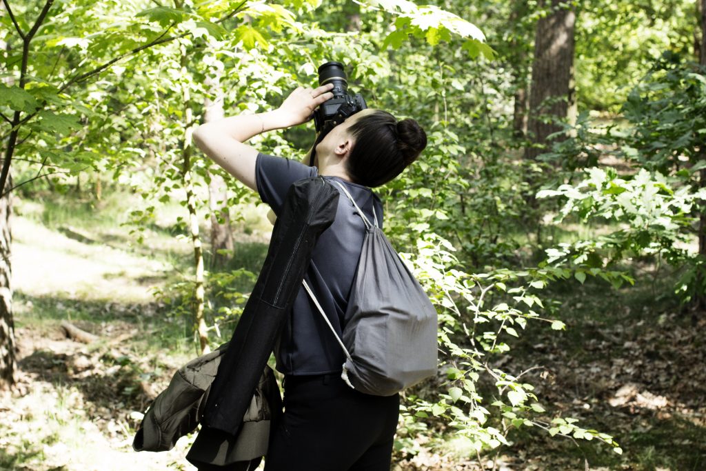Eine junge Frau schaut durch eine Kamera nach oben. Sie steht in einem Wald, zu sehen sind Äste und Büsche mit grünem Laub. Die Frau trägt einen Rucksack sowie eine Umhängetasche. Sie sieht schwer bepackt aus, ist aber sehr konzentriert und fokussiert auf ihren Blick durch die Kamera.