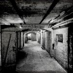 Der schmale Flur eines Kellers ist auf einem schwarz-weiß Foto zu sehen. Das pure Mauerwerk untersetzt mit Türen. Der Flur wird in der Flucht es Bildes schmaler.